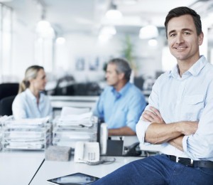 Confident businessman sitting on a desk with his coworkers in the background